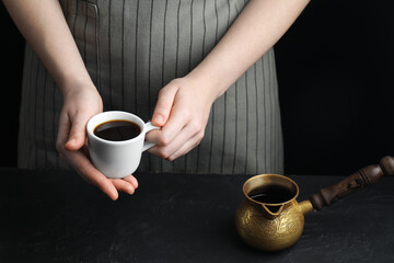Turkish coffee. Woman with cup of freshly brewed beverage and cezve at black table, closeup