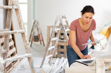 Naklejka premium Asian woman construction manager checking project documentation and using laptop while working in apartment.