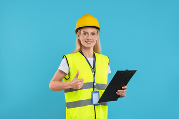 Engineer in hard hat holding clipboard and showing thumb up on light blue background