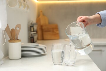 Woman pouring water from jug into glass at white table in kitchen, closeup. Space for text