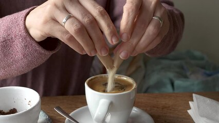 A close-up video showing a woman tearing open a brown sugar paper stick and sweetening her coffee in a white espresso cup, capturing a cozy, homemade beverage moment.