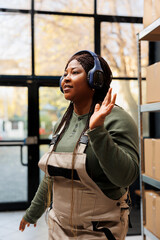 Cheerful manager wearing headphones listening music during work break in storehouse, dancing and...