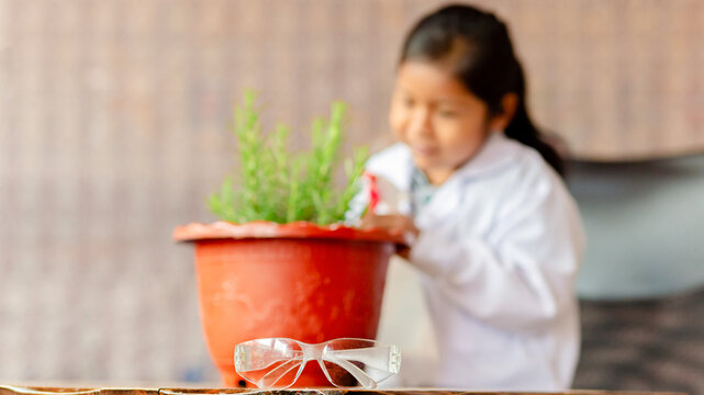 Little Latina girl in a biologist's coat and safety glasses, watering and observing plants in her pots, concept of saving the world, ecological awareness, and environmental learning.
