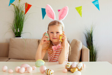 Blond child girl wearing bunny ears on Easter day play with Easter eggs at the table at home