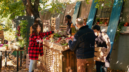Naklejka premium Young farmer serving client with various produce and selling homegrown farm veggies at local food market. Female customer buying fresh organic fruits and vegetables, farming nutrition.
