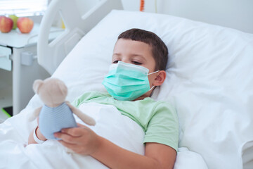 Portrait of a cute European seven year old boy wearing a medical mask and a light green T-shirt lying on a bed in a hospital room and playing with his teddy bear - 728134659