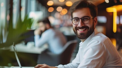 Obraz premium cheerful businessman in eyeglasses typing on laptop while sitting near colleague with mobile phone and bearded man on blurred foreground in lounge of modern coworking space