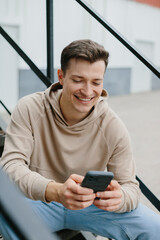 A young man is sitting on the metal stairs on the street, holding the phone in his hands.