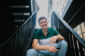 Cheerful young man sitting on the stairs on the street, looking at the camera.