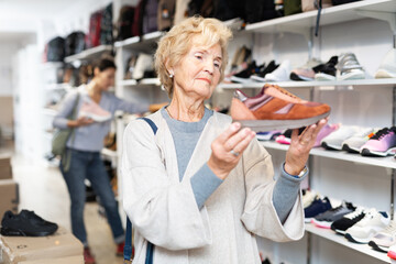 Fototapeta na wymiar Mature european woman who came to a shoe store for shopping, chooses sports sneakers, standing near the shelves with the goods
