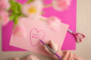DIY mom card. Mothers Day.Child draws a heart on a pink piece of paper and writes mom I love you....