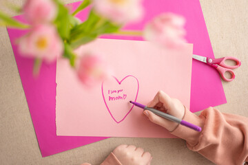 moms day.DIY mom card.Mothers Day.Child draws a heart on a pink piece of paper and writes mom I...