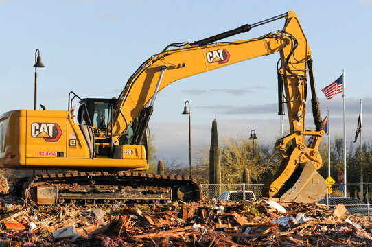 A large CAT hydraulic excavator machine sits atop the rubble of a demolished building, with an American flag waving in the background