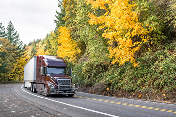 Shiny brown big rig semi truck with dry van semi trailer driving on the winding narrow road with autumn trees on the hillsides