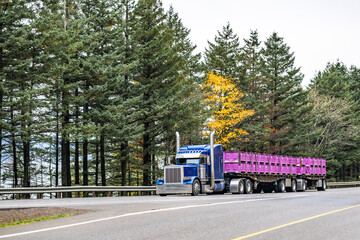 Classic big rig blue semi truck tractor transporting loaded boxes fastened on two flat bed semi trailers running on the autumn highway road