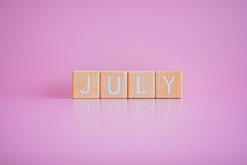 Wooden blocks form the text "JULY" against a pink background.