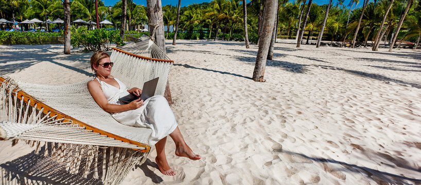 Woman relaxing on a hammock with a laptop on a sunny beach, concept of remote work or digital nomad lifestyle, background with a place for text - Powered by Adobe