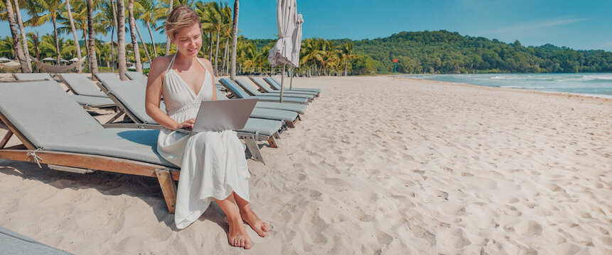 A woman works on a laptop while sitting on a beach chair against a beautiful tropical beach backdrop, illustrating the concept of remote work or digital nomad lifestyle