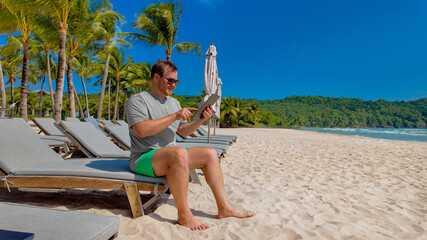 Man in casual summer clothing sitting on a beach chair and reading a tablet on a sunny tropical...