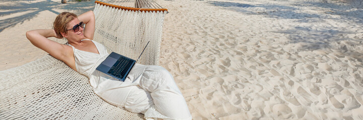 A person relaxing on a hammock with a laptop on a sandy beach, illustrating remote work or a...