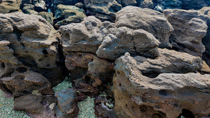 Rugged, weathered volcanic rocks with holes and crevices near a coastline, depicting natural geological textures and formations