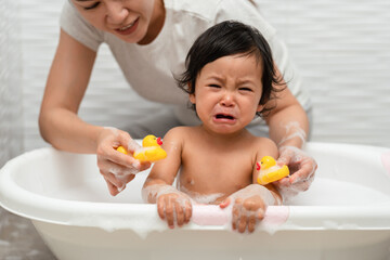 mother bathing her crying infant baby with foam bubbles in bathtub