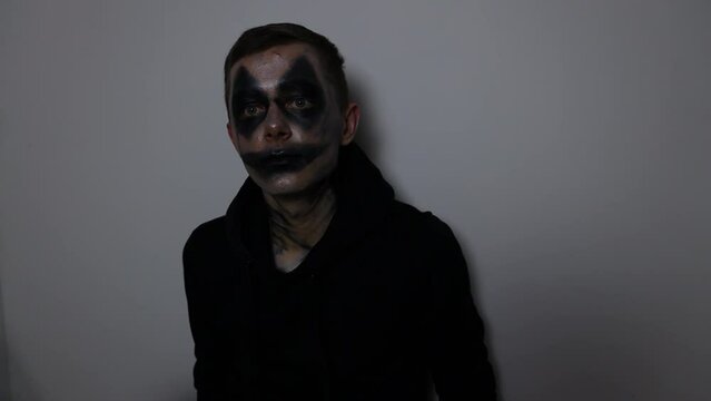 A man with a black clown makeup mask sits down at the floor and stares at the camera