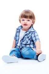 Smiling little boy 2 years old sitting on the floor. Child in a plaid shirt and jeans. Happy childhood. White background. Vertical.
