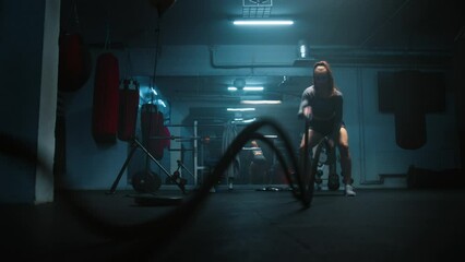 Female athlete exercises with battle ropes in dark boxing gym with LED lighting. Female boxer does cardio or endurance workout before championship fight. Physical activity and CrossFit training. - Powered by Adobe