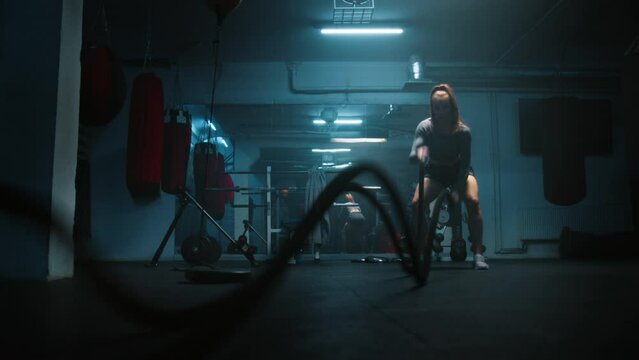 Female athlete exercises with battle ropes in dark boxing gym with LED lighting. Female boxer does cardio or endurance workout before championship fight. Physical activity and CrossFit training.