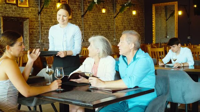 Adult family of three is relaxing in cafe. Waitress girl serves glass of wine and helps visitors decide to choose snack on menu