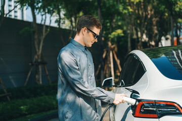 Young man travel with EV electric car charging in green sustainable city outdoor garden in summer...