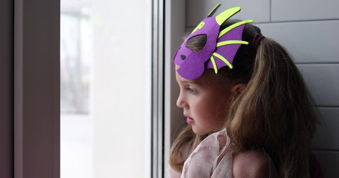 Girl with dragon mask on forehead looks out apartment window watching passers-by in big city. Suit for children to play and improve imagination