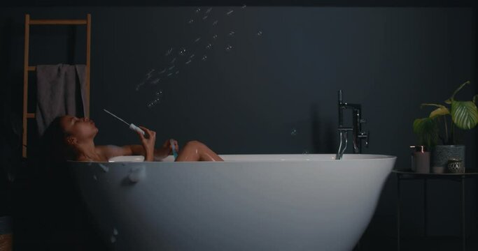 Side view happy African American young female blowing on soapy foam lying in stylish modern bath.