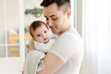 Side view of smiling young crop father in casual clothes looking down while embracing cute little...