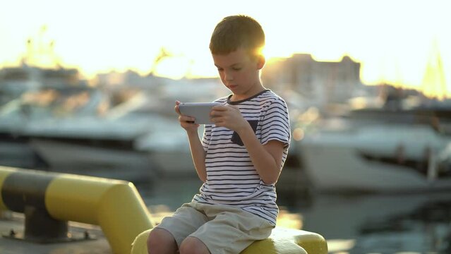An eight-year-old boy sits on the shore and plays the phone against the backdrop of yachts and boats.