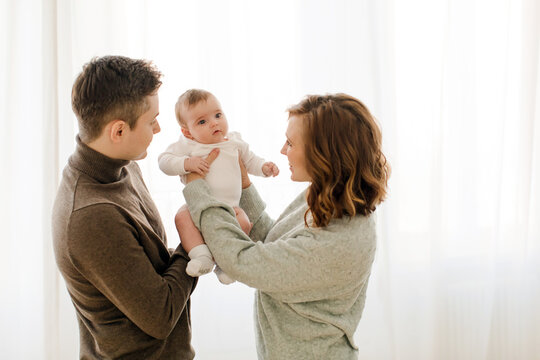 Side view of cheerful young couple looking away while carrying baby in hands with socks in living room against curtain with reflecting sunny daylight