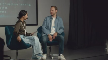Full shot of cheerful Caucasian male CEO of tech startup and Asian female moderator sitting together on stage at IT conference, woman with headset asking questions, both joking and laughing