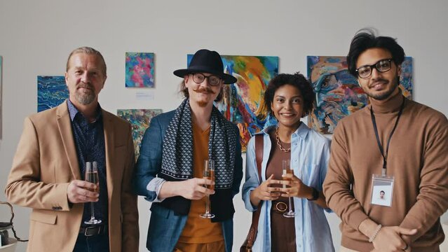 Medium portrait shot of multiethnic modern art museum employees and guests standing in front of paintings at exhibition opening, with champagne, discussing artworks, looking at camera and smiling