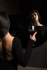 Luxurious woman in a black dress posing with a glass of wine near the mirror