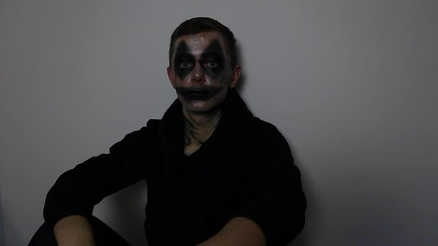 Young Caucasian man with a scary dark clown Halloween costume sitting down wearing a black hoodie