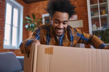 Fotobehang Low angle view of a cheerful multiracial young man at home opening a cardboard box and putting out a cobalt blue running long sleeve top mens.  © Straxer