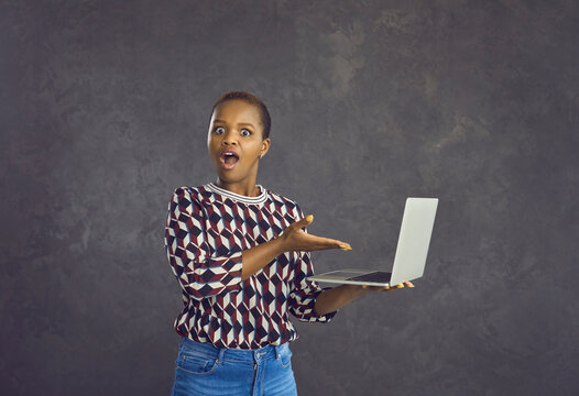 Portrait of stunned gen z African American girl on black studio background shocked by sale offer or discount online on laptop. Amazed young black woman surprised by unexpected news on computer.