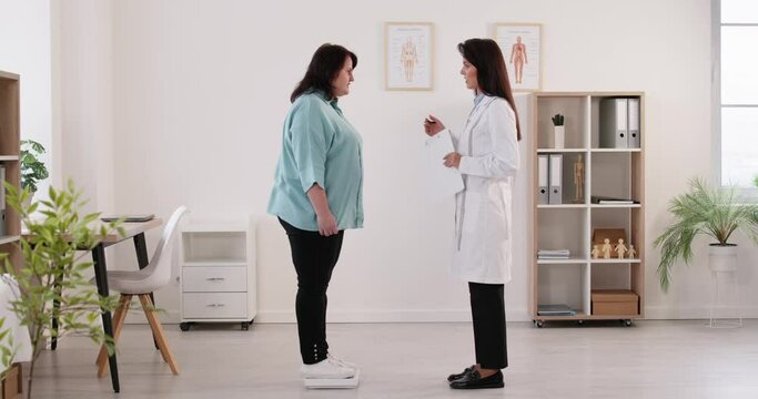Doctor nutritionist measuring weight on scales of overweight woman in clinic and writing down results. Fat patient having consultation with physician about weight loss and diet in office. 4k video.