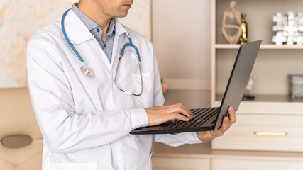 Serious young professional male doctor using laptop computer in hospital office watching medical webinar training, writing in healthcare report, consulting patient online at telemedicine meeting.