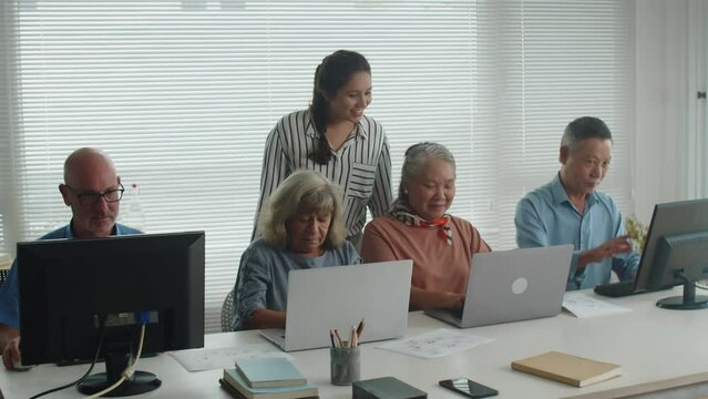 Wide shot of young female teacher checking diverse senior students progress during computer class