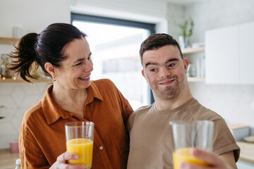 Portrait of young man with Down syndrome with his mother at home, toasting with juice. Morning routine for man with Down syndrome. - obrazy, fototapety, plakaty