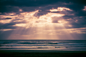 Sunset over Muriwai beach. Sun beams breaking through heavy clouds as waves splashing into wet...