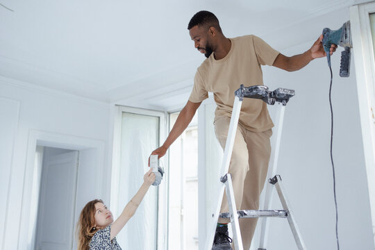 Woman giving ear muff to man standing on ladder and painting wall