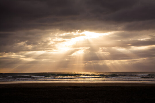 Sunset over Muriwai beach. Sun beams breaking through heavy clouds as waves splashing into wet sand. Auckland, New Zealand